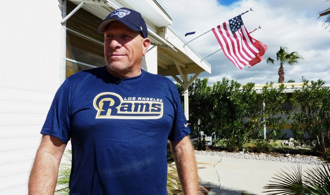 A man wearing a Los Angeles Rams t-shirt and a cap standing outdoors near a house with an American flag waving in the background.