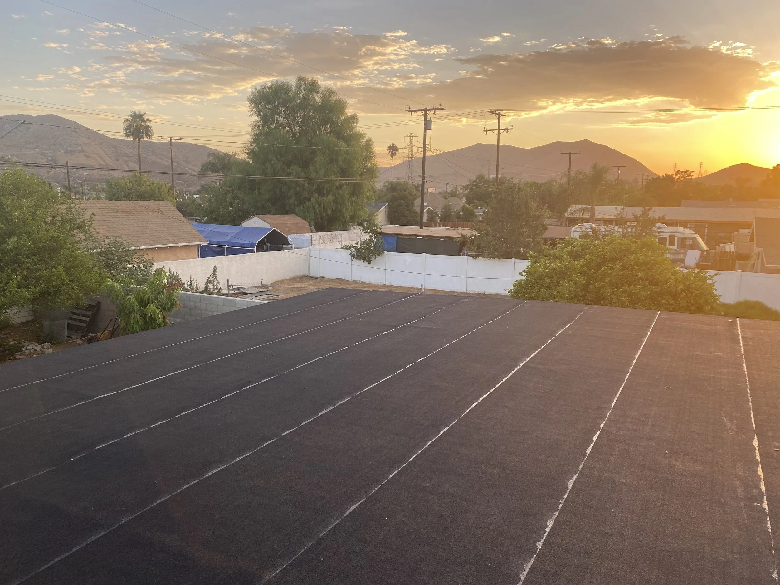 View of a neighborhood at sunset with mountains in the background, trees, power lines, and rooftops, with the foreground showing a flat rooftop with a black sunshade or screen.