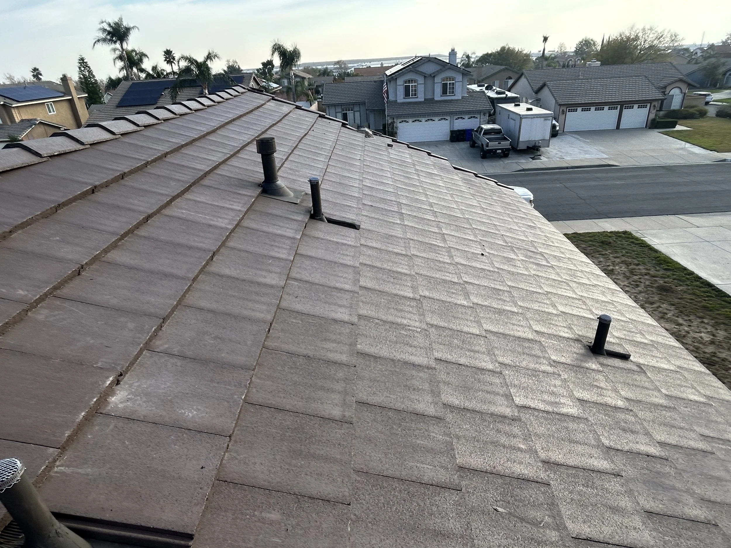 View of residential rooftop with vents and neighboring houses, palm trees, street, and parked vehicles in the background.