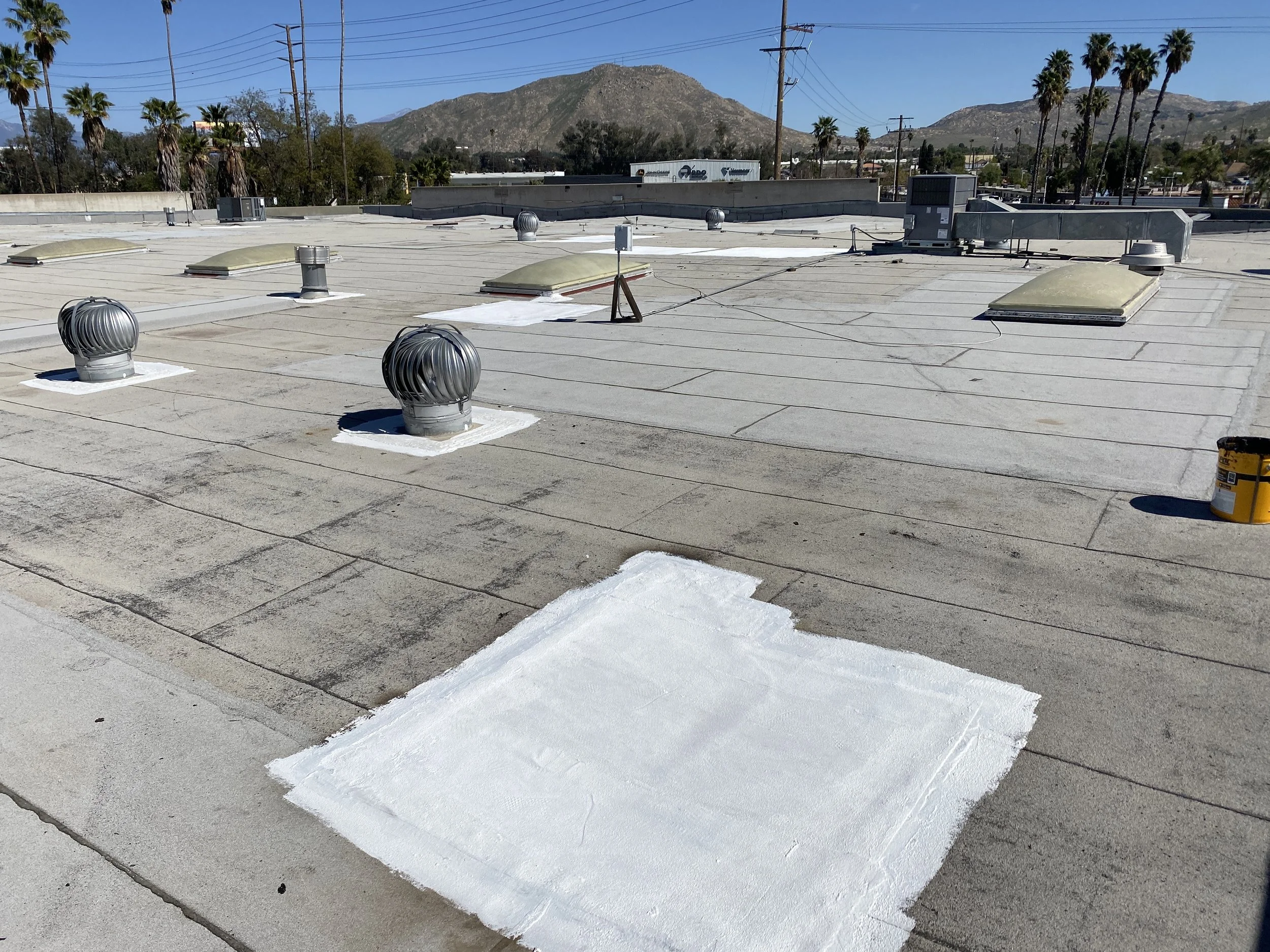 Flat rooftop with vents, skylights, and HVAC units under a clear blue sky, with mountains and palm trees in the background.