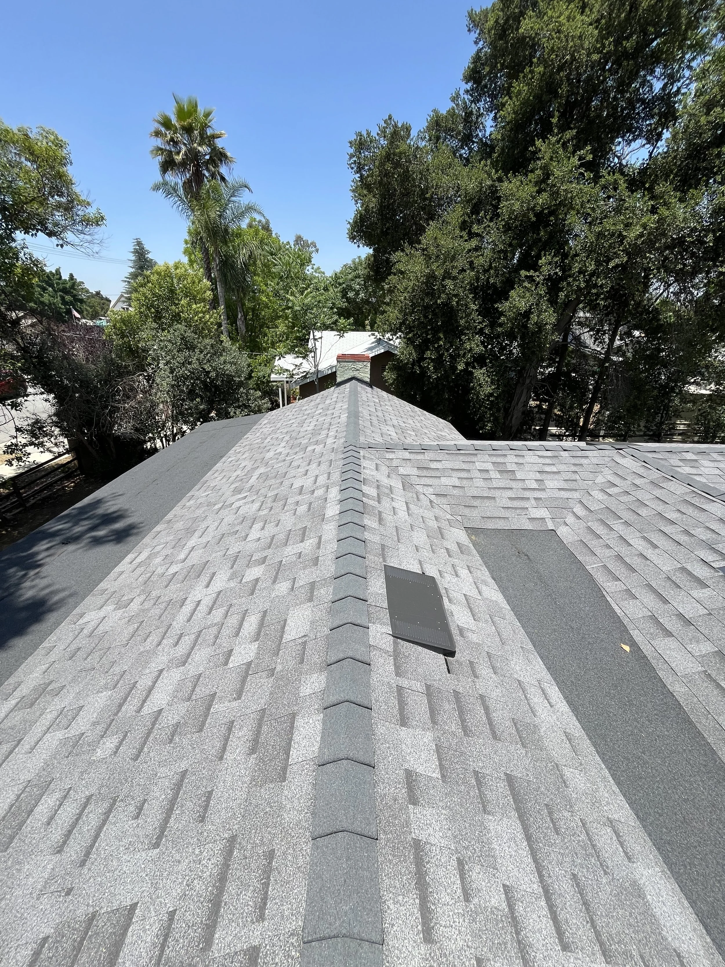 View of a house rooftop with gray shingles, a solar panel, and surrounding trees under a clear blue sky.