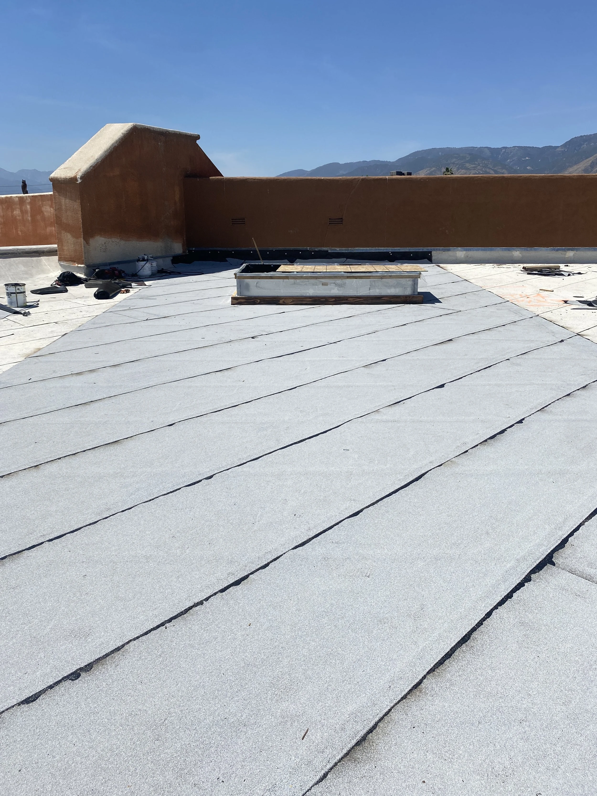 Flat rooftop with gray roofing material, a vent pipe in the middle, a brown wall with a small chimney, and mountains in the background under a clear blue sky.