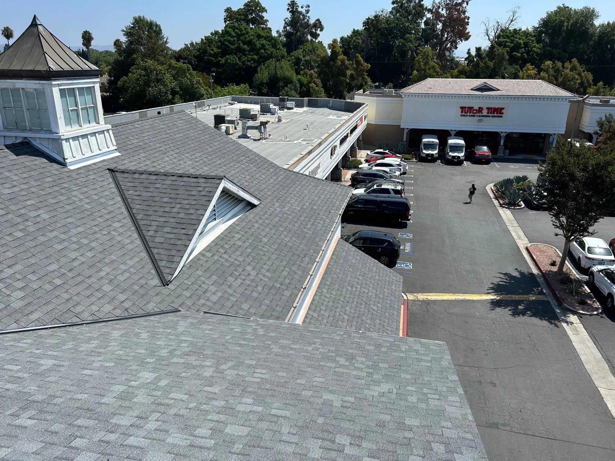 Aerial view of a shopping center parking lot with several cars parked and a person walking. In the background, a store named 'TUTOR TIME', with a white exterior, is visible along with trees and a clear sky.