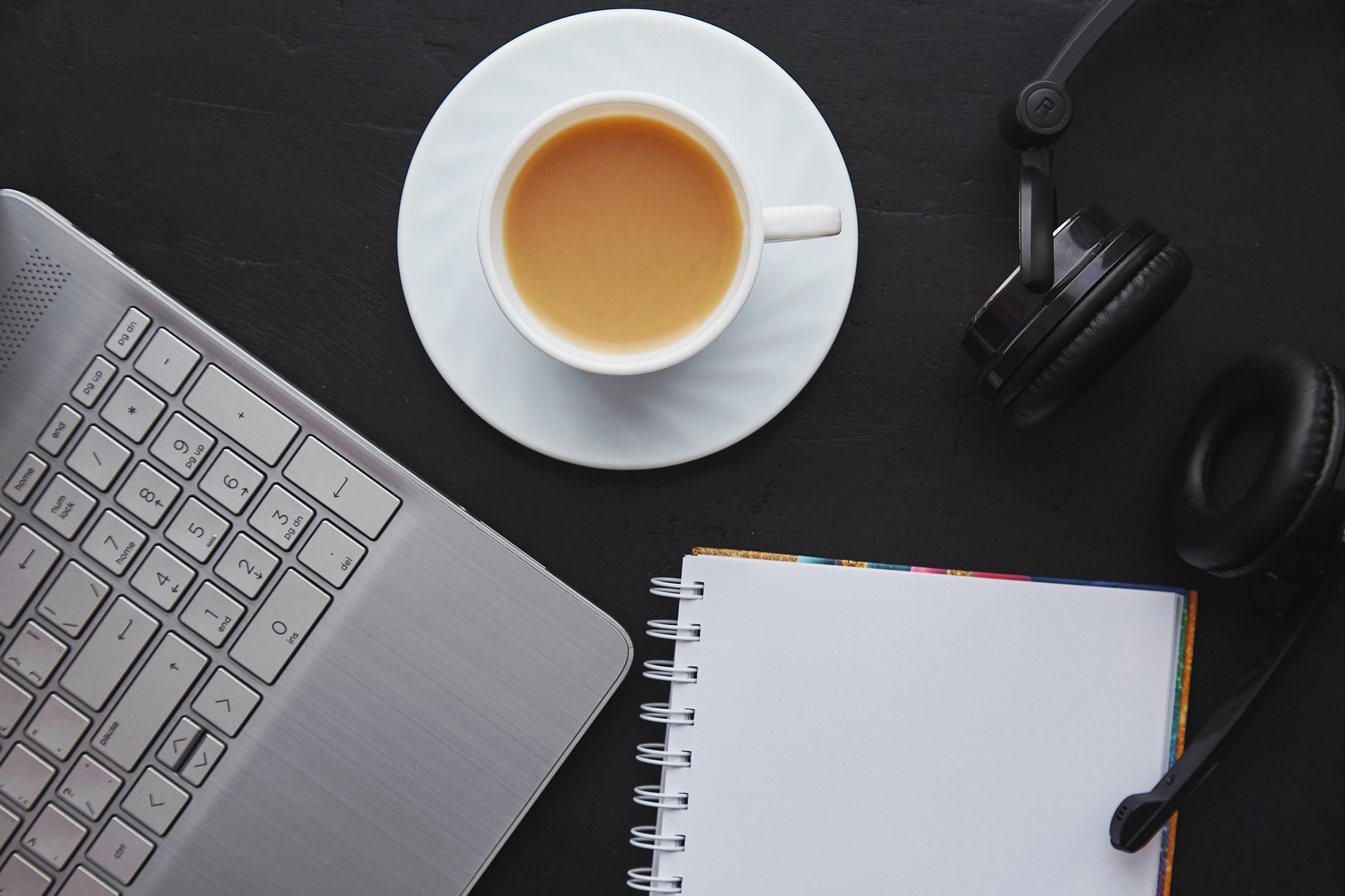 Top view of a workspace with a silver laptop, a white cup of coffee on a saucer, a pair of black headphones, a pen, and a spiral notebook on a black surface.