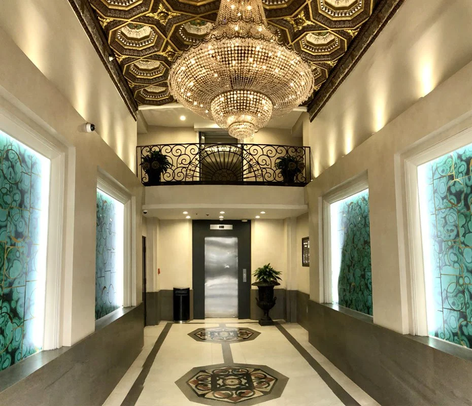Elegant hotel lobby with a large chandelier, decorative ceiling, elevator, potted plant, and illuminated stained glass windows.