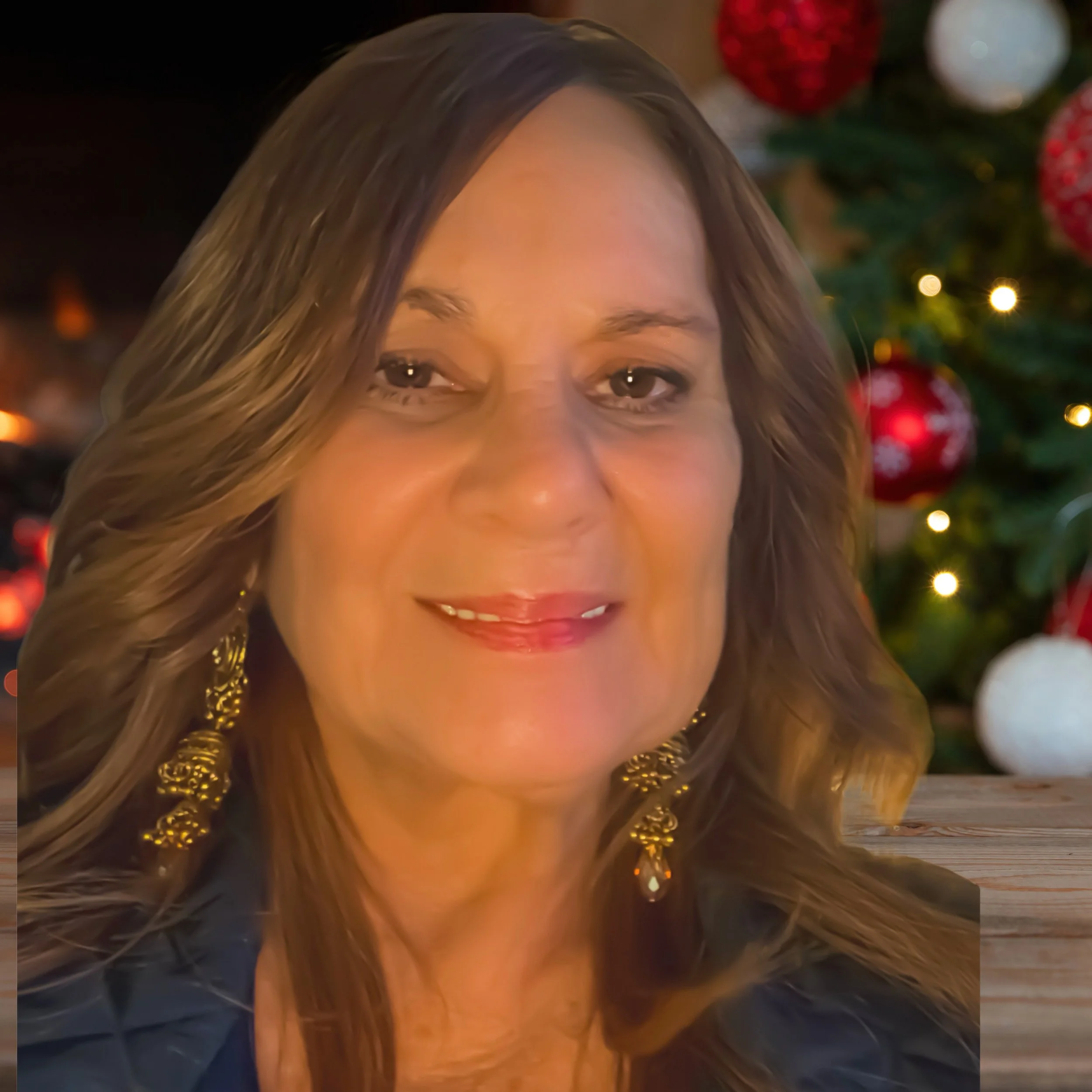 Denise@dp-artwork.com Close-up of a smiling woman with long brown hair and gold earrings, with a decorated Christmas tree in the background.