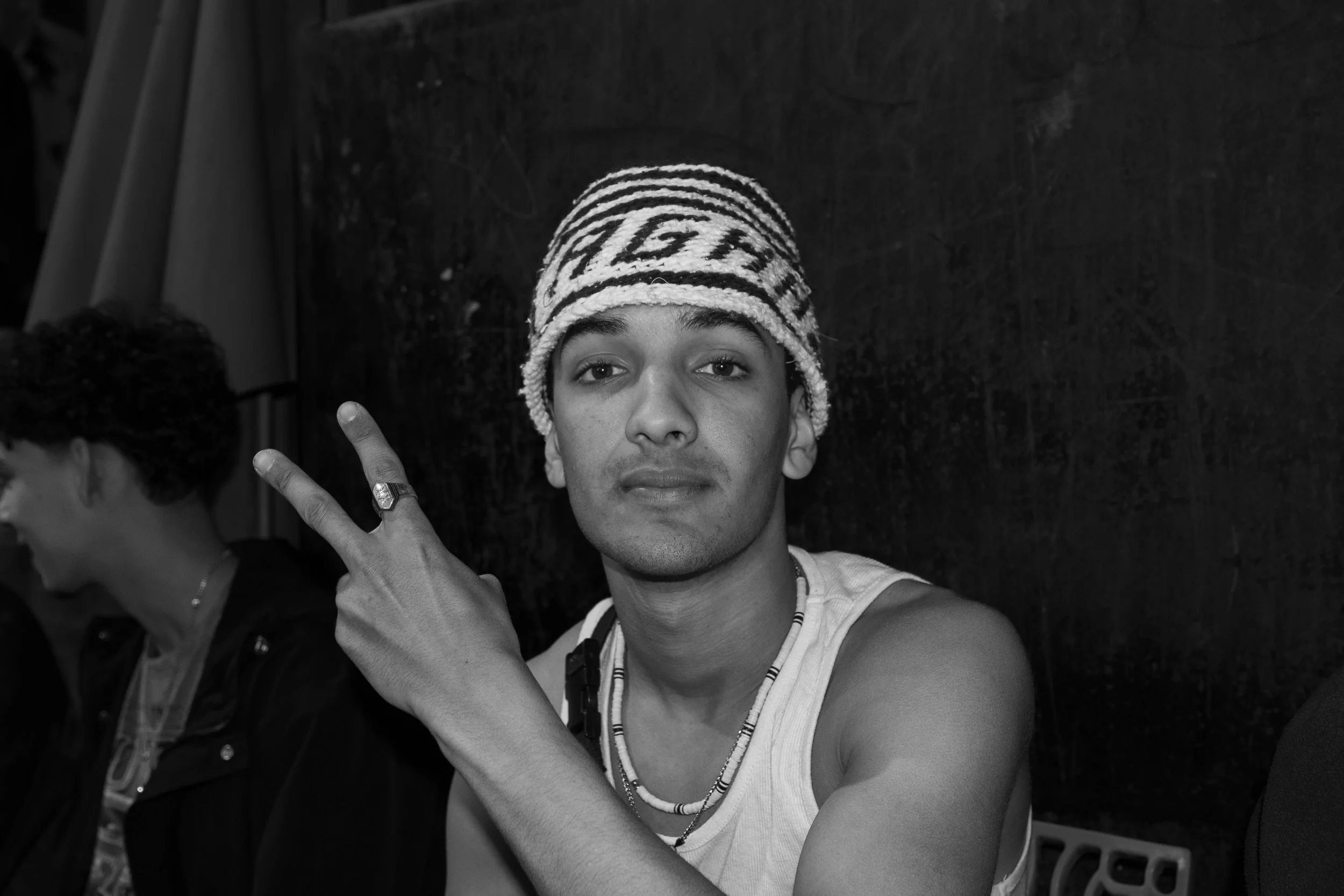 Young man with a patterned beanie making a peace sign gesture, wearing a sleeveless shirt and layered necklaces, sitting against a dark background, with another person partially visible on the side.