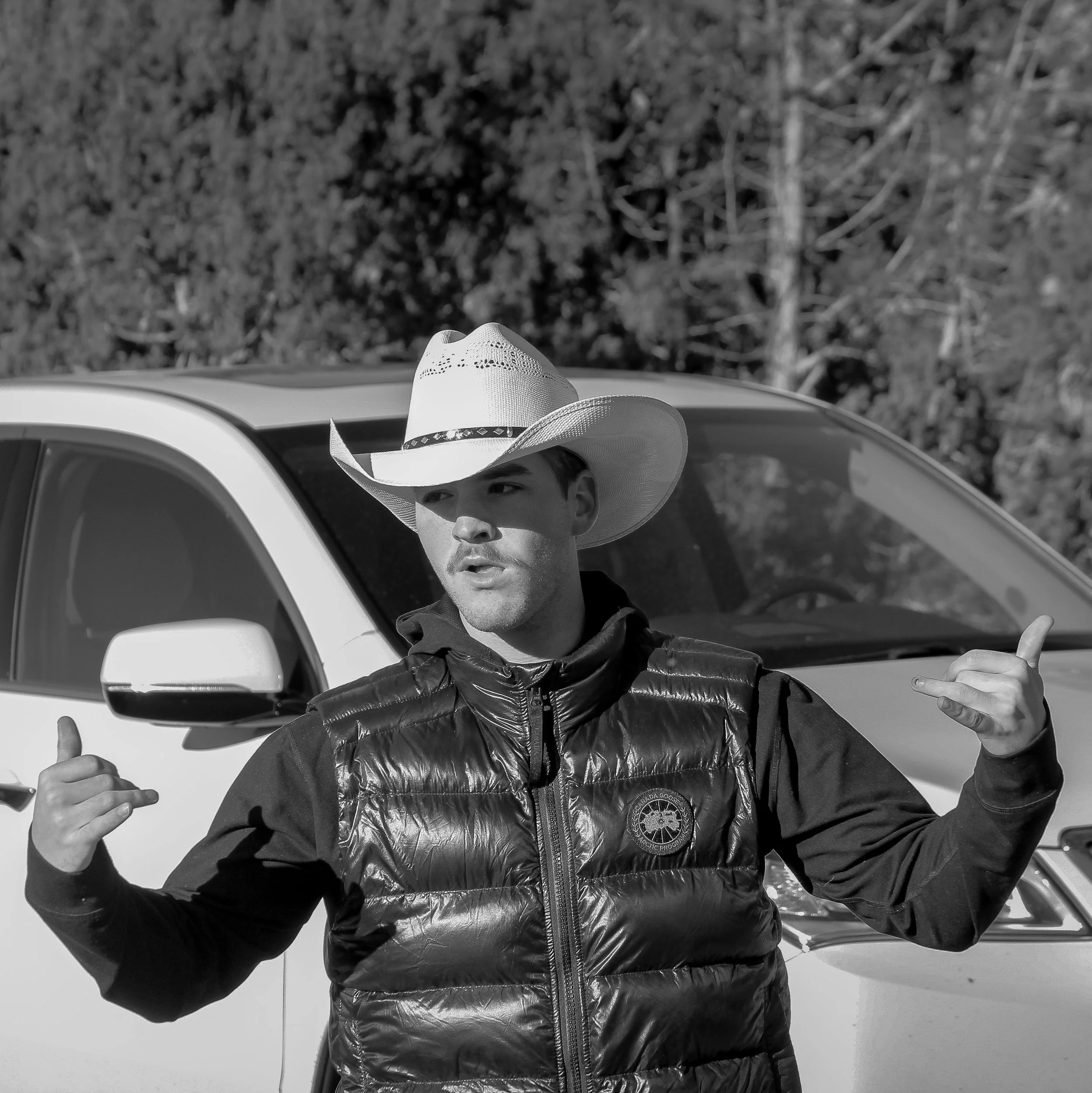 A man in a cowboy hat and black puffy vest making gesture with both hands, standing in front of a white vehicle outdoors.