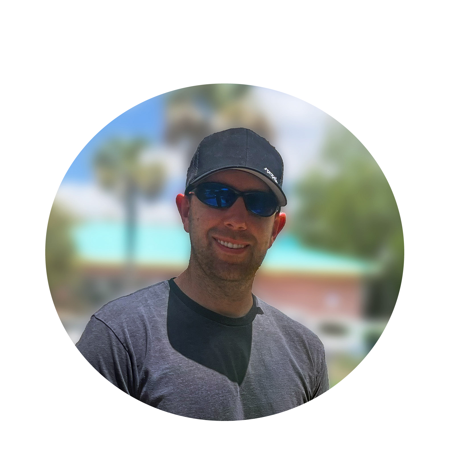 A smiling man wearing sunglasses and a black cap outdoors on a sunny day with palm trees and a building in the background.