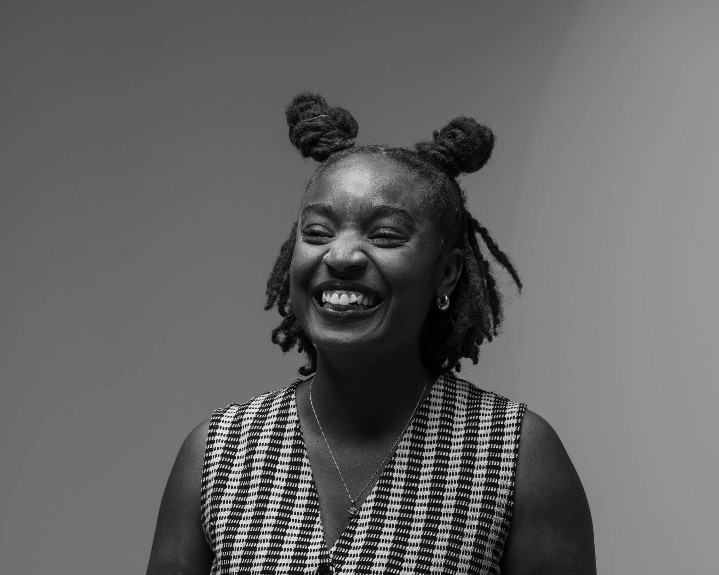 Black and white photo of a smiling woman with curly hair styled in two buns and wearing a patterned sleeveless top, with a plain background.