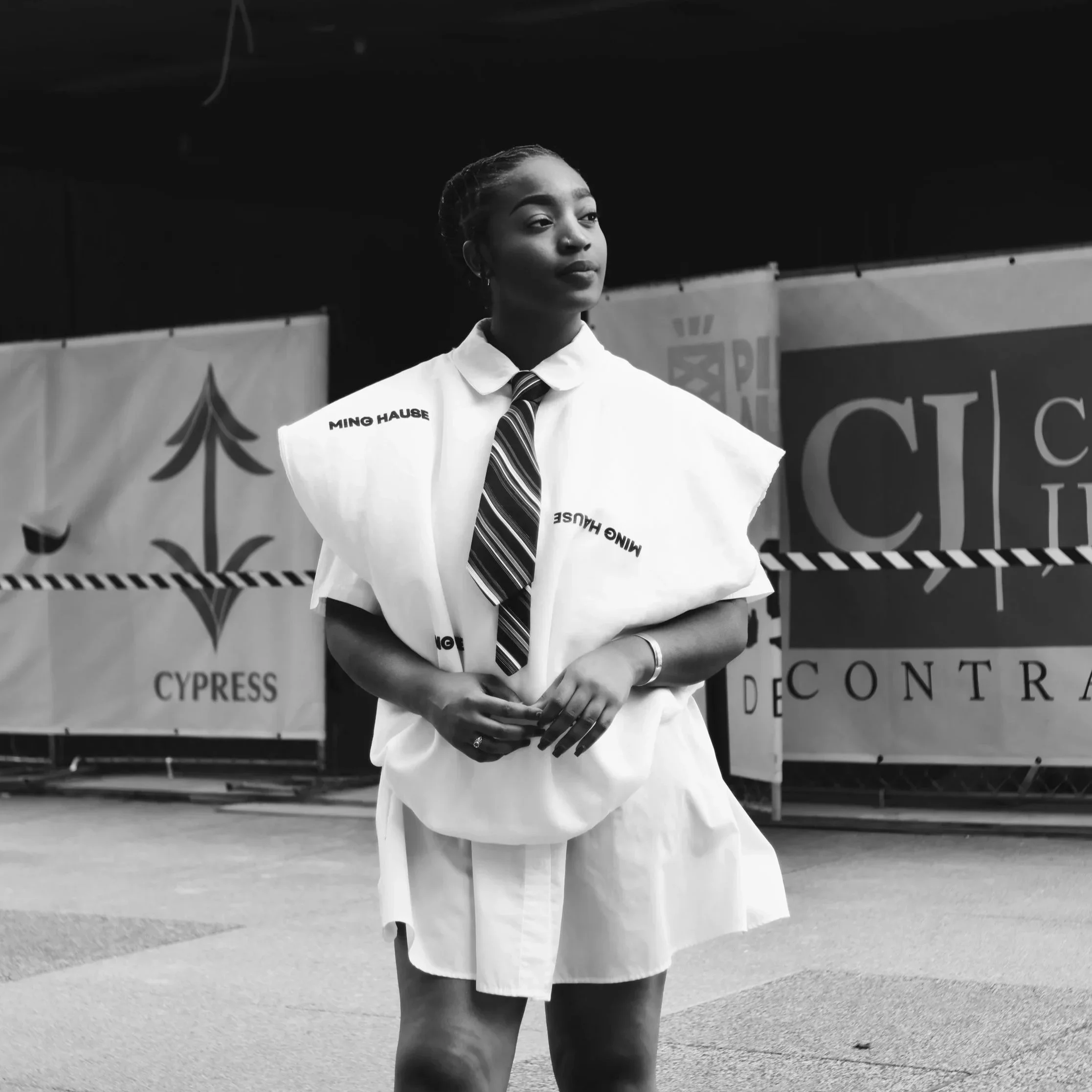 A young woman stands confidently with arms crossed, wearing a white oversized shirt with bent sleeves, a striped tie, and a skirt. She has braids and a serious expression, standing in front of banners that say 'Cypress' and other text, at an indoor event or rally.