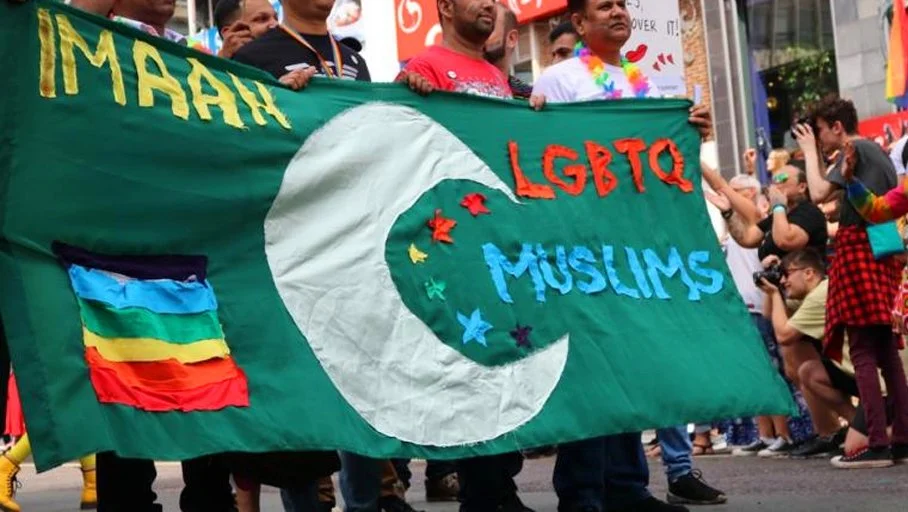 A group of people participating in a pride parade holding a large banner with the words 'IMAN', 'LGBTQ', and 'MUSLIMS'. The banner features a rainbow flag and a crescent moon symbol.