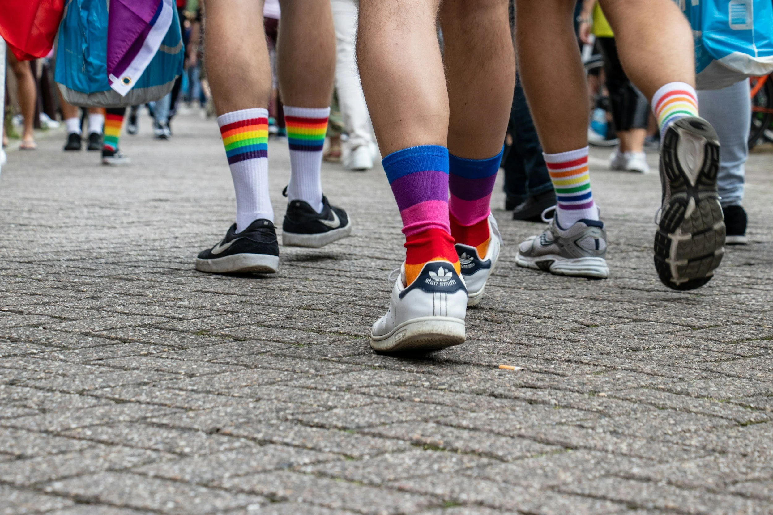 People walking outdoors on a paved surface, wearing colorful rainbow-striped socks and casual shoes, with a crowd in the background.