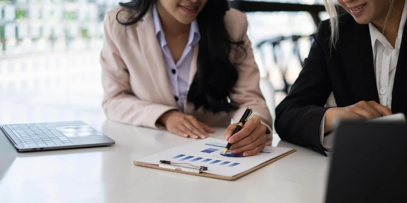 Two women in business attire reviewing charts and graphs at a meeting, with a laptop and tablet on the table.