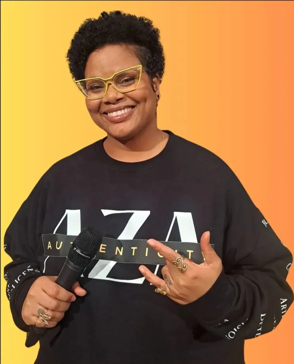 A smiling woman with short curly hair and glasses holding a microphone against a yellow and orange background, wearing a black sweatshirt with white letters and text.