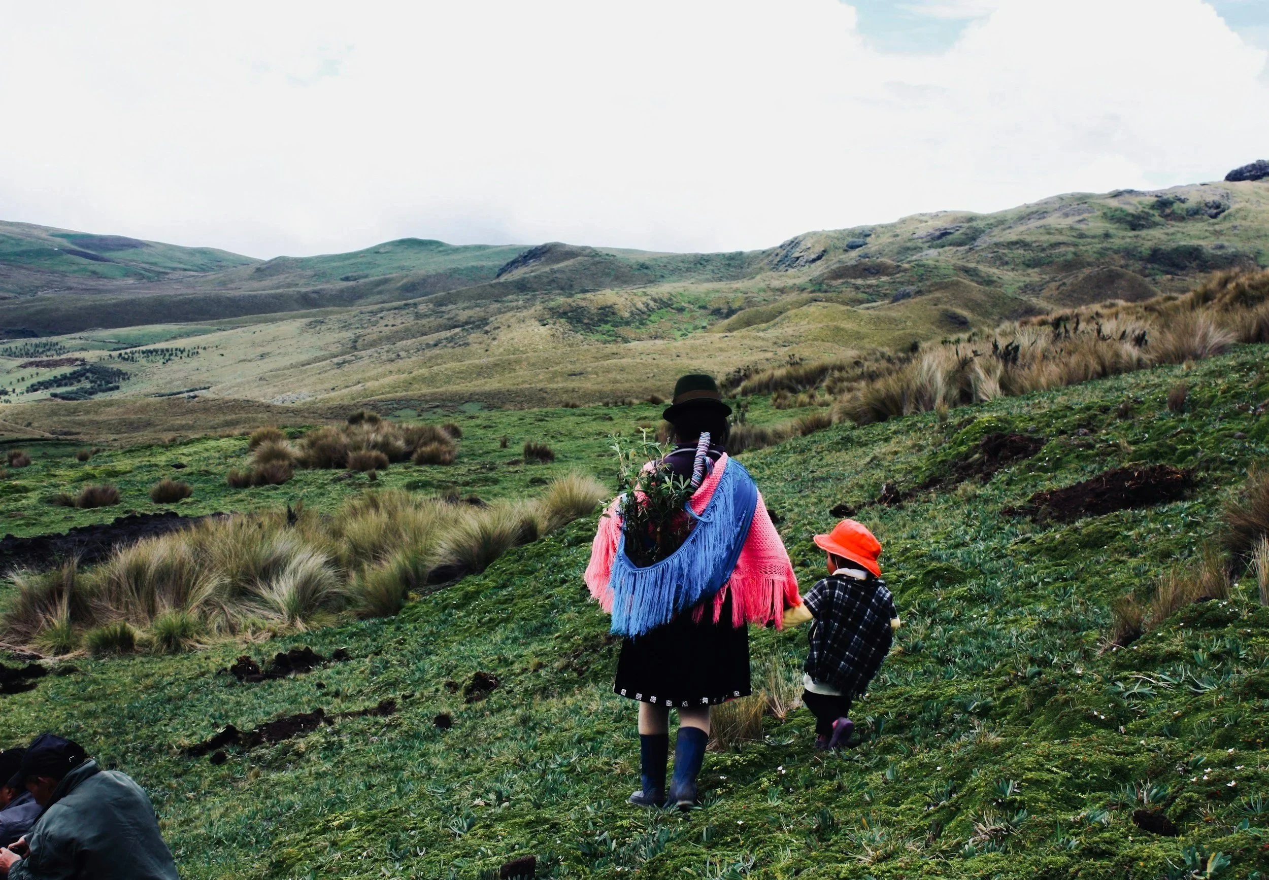 A woman and a young child walking through a green, hilly landscape with tall grass and sparse vegetation, under a cloudy sky.