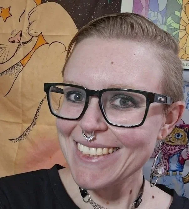 A smiling person with short blonde hair wearing large black glasses, a septum ring, and a choker necklace, standing in front of colorful illustrated posters.