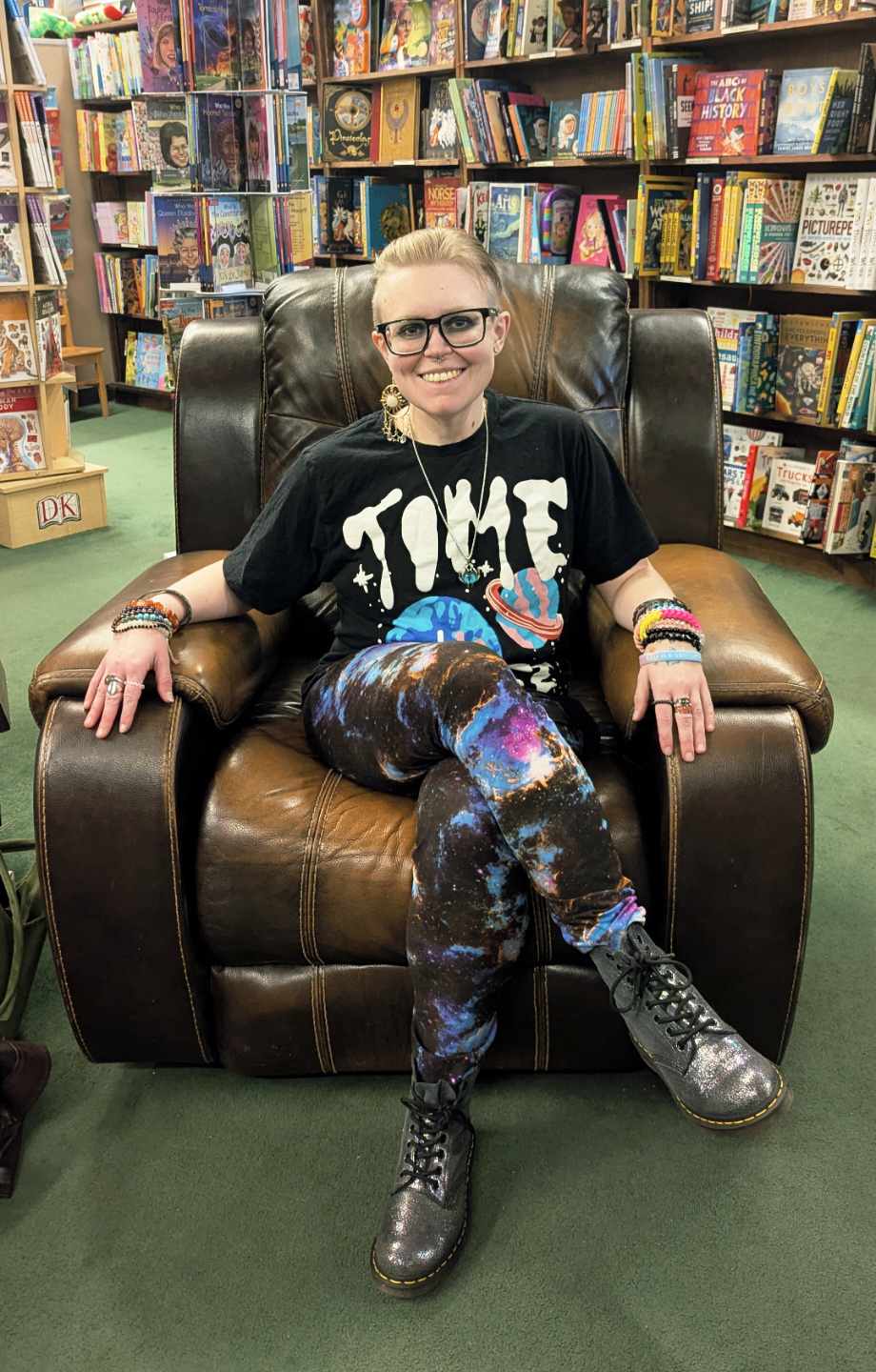 An image of Sage Nestler, MSW, a white person with glasses and blonde hair.  They are sitting in a chair with bookshelves behind them, wearing galazy leggings and a space themes shirt with multiple rings, bracelets, necklaces, and earrings.