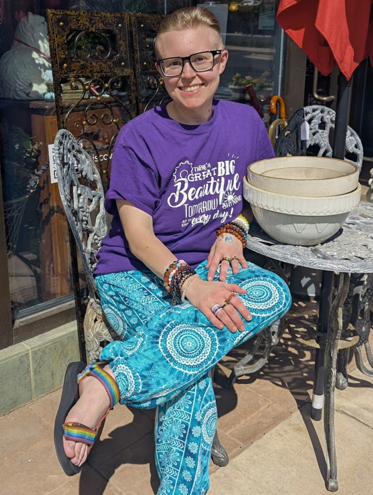 Person sitting on a decorative outdoor metal chair, smiling, wearing a purple T-shirt with white text, blue patterned pants, rainbow sandals, and rainbow bracelets. They are sitting with one leg crossed over the other, in front of a table with large ceramic bowls, outside a shop with glass windows.