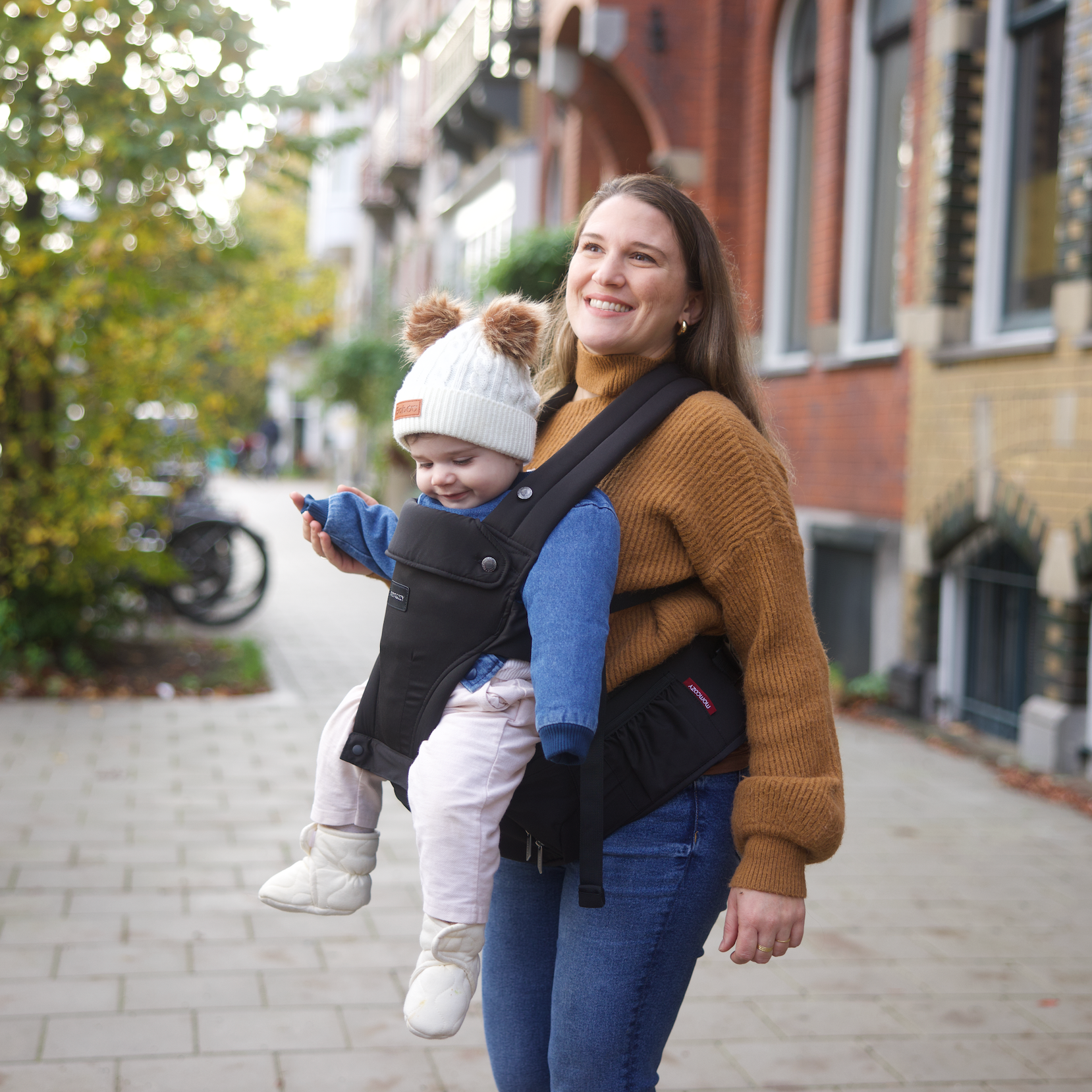 A woman smiling and walking outdoors with a child in a baby carrier on her chest. The child is dressed in a white hat with bear ears, blue jacket, and white pants. The background shows a sidewalk, greenery, and residential buildings.