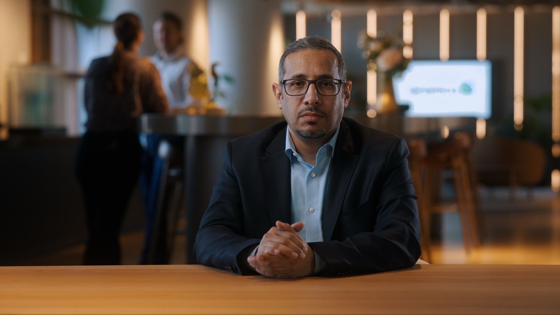 A man in glasses and a business suit sitting at a wooden table in a modern, professional-looking office or meeting space. Two people are blurred in the background, engaged in conversation, with a computer monitor and decorative plants visible.