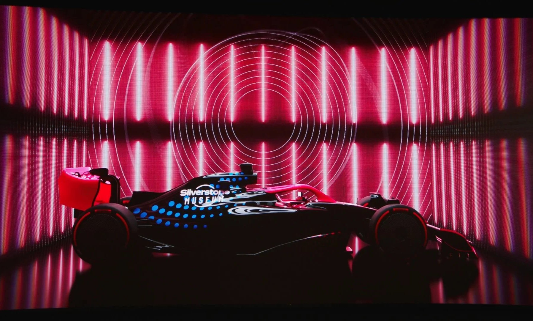 A futuristic racing car with a black and red design at a museum exhibit, illuminated by neon pink and purple lights with circular patterns in the background.