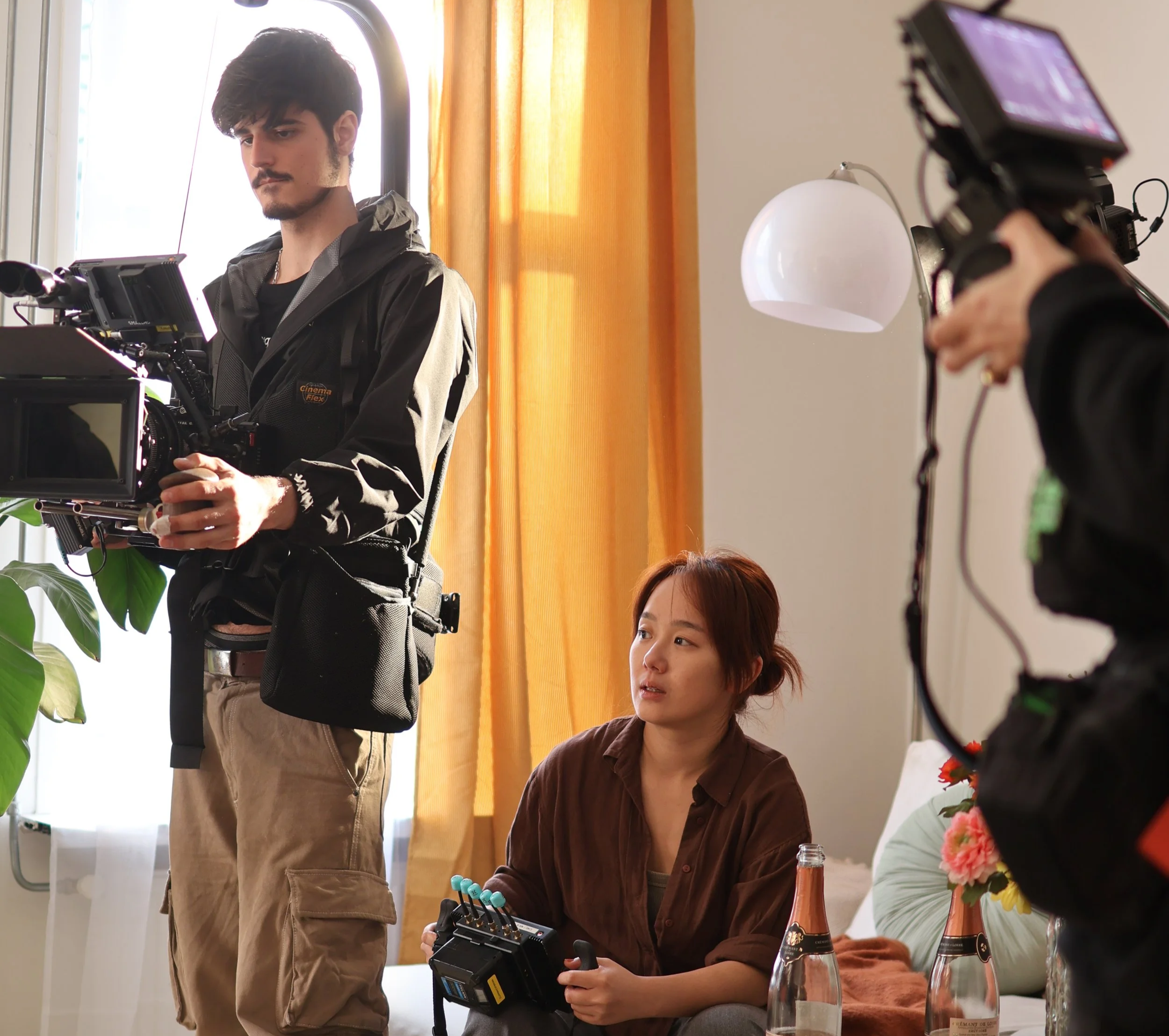 A film crew shooting a scene indoors with a female actress sitting on a couch, surrounded by filming equipment, with a camera operator and a director or crew member nearby.