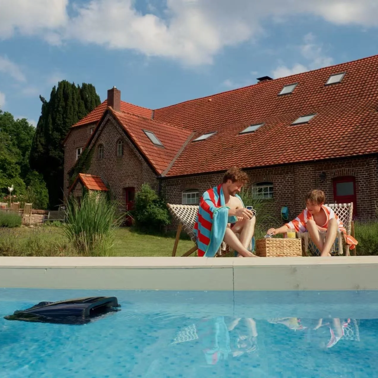 Two people by a swimming pool with a house and red-tiled roof in background. One is sitting on a lounge chair, the other on the ground, both wearing beach towels and playing with phones. A robotic pool cleaner is in the pool.