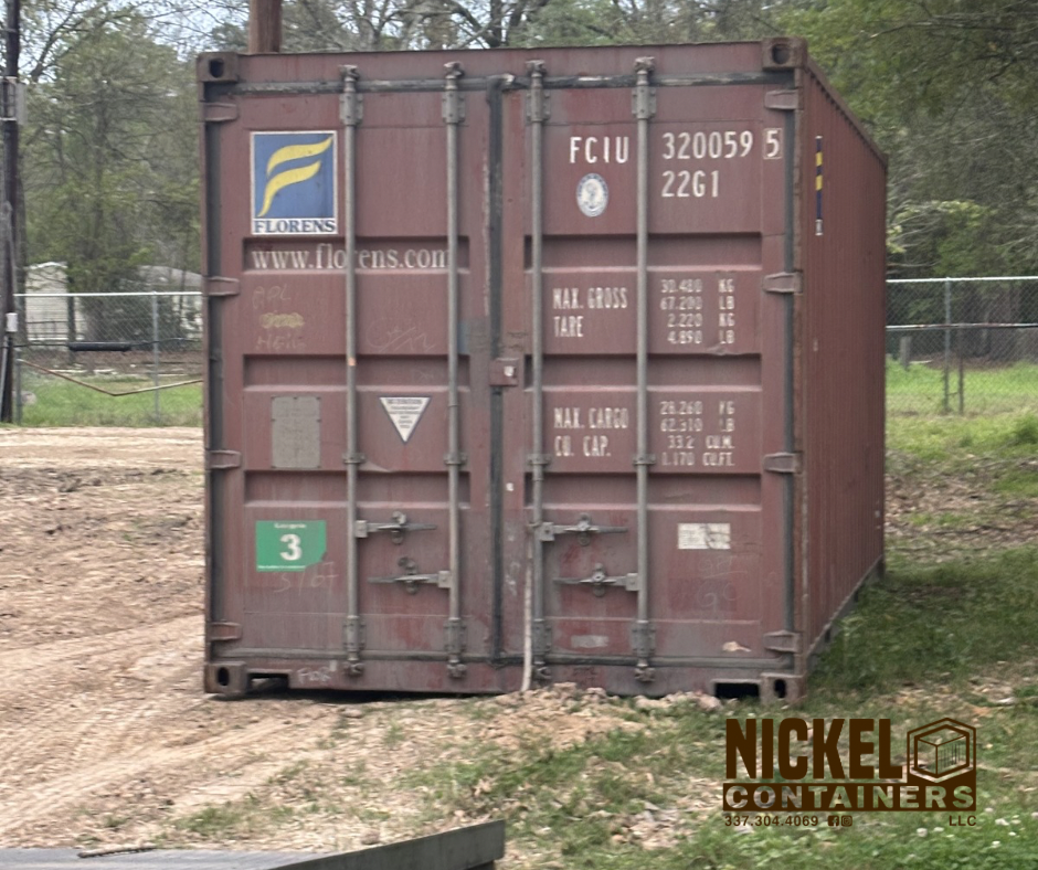 A red shipping container with various markings, located outdoors on a dirt ground with grass and trees in the background.