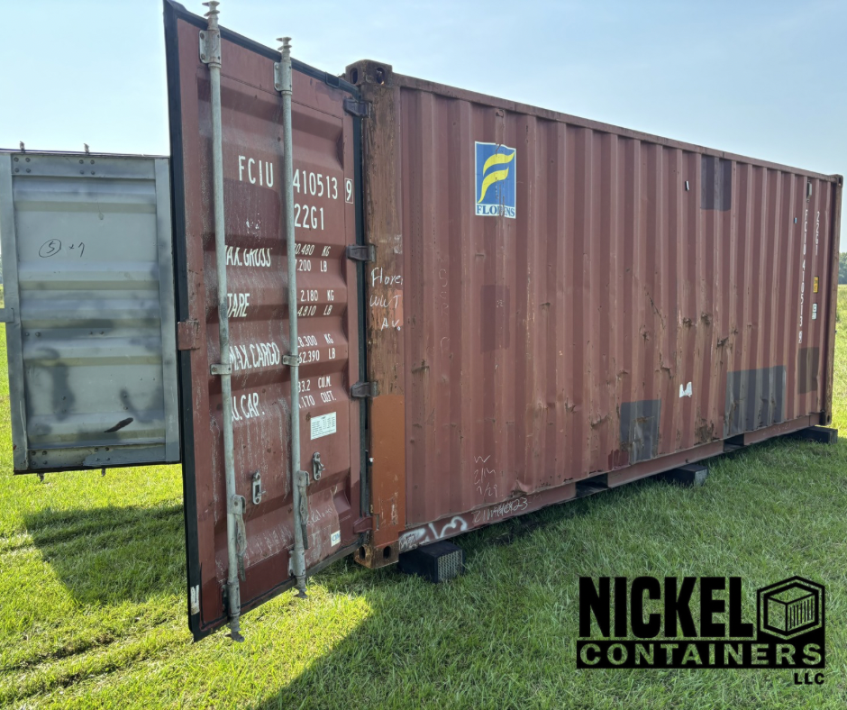 A large, rusty shipping container with a small gray container attached on the left side, situated on grass under a cloudy sky. There is graffiti on the container walls, a logo on the upper right, and text markings on the doors.