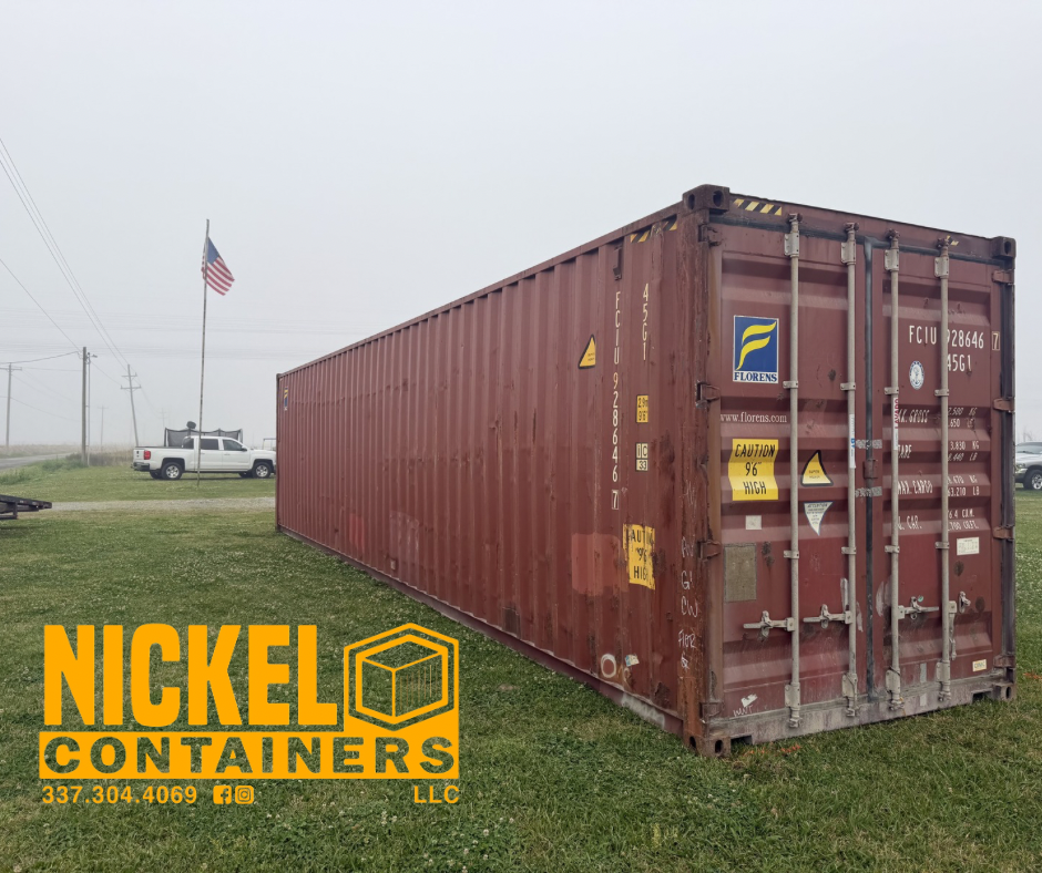 A large red shipping container with multiple warning labels, placed outdoors on grassy land. There's a white pickup truck and an American flag on a pole in the background, along with power lines and overcast sky.