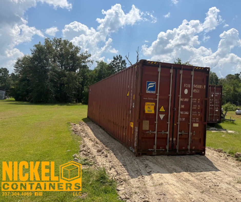 Red shipping container on dirt path in a grassy area with trees and a partly cloudy sky overhead.