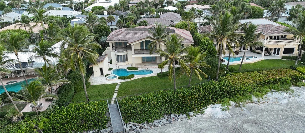 Aerial view of beachfront houses with pools, palm trees, lush green lawns, and a sandy beach shoreline.