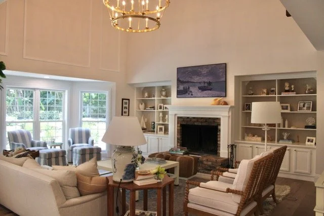 Living room with cream sofas, armchairs with striped upholstery, cream armchairs, and wooden side tables. Fireplace with a painting above it, built-in shelves on either side displaying photos and decor. Large windows with white framing and a chandeli