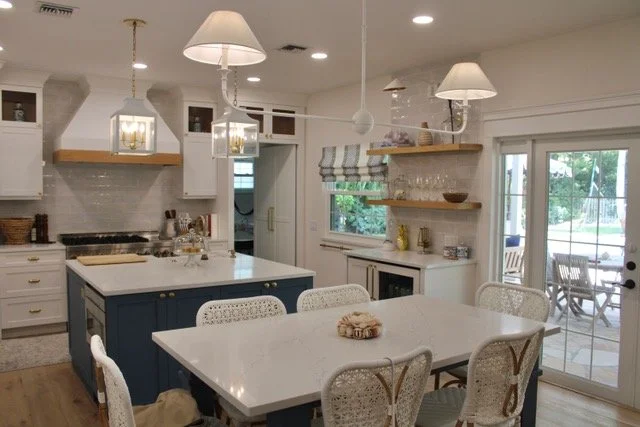 Kitchen with white cabinets, a blue island, and a white dining table with six chairs; pendant lights and open shelves on the wall; sliding glass door leading to an outdoor patio.