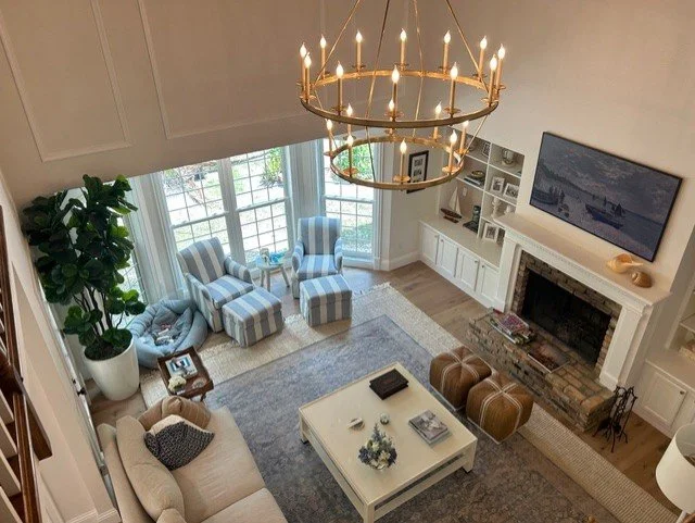Living room with large windows, a chandelier, a sofa, striped armchairs, a coffee table with decor, a fireplace with a TV above, built-in shelves, and a potted plant.