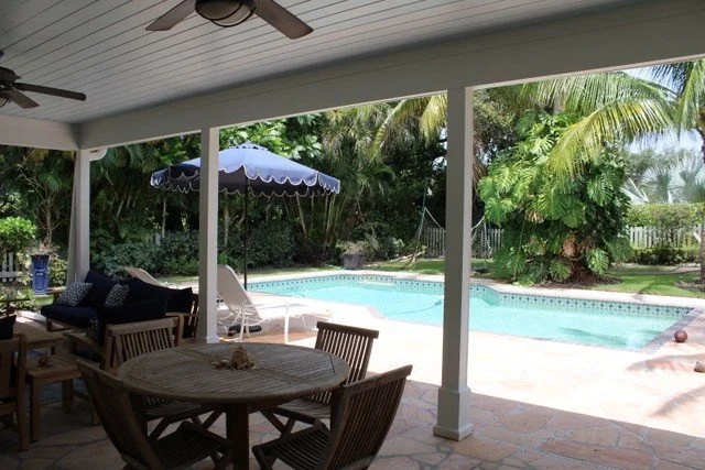 Covered patio overlooking a backyard swimming pool, surrounded by lush palm trees and greenery, with outdoor furniture and an umbrella.