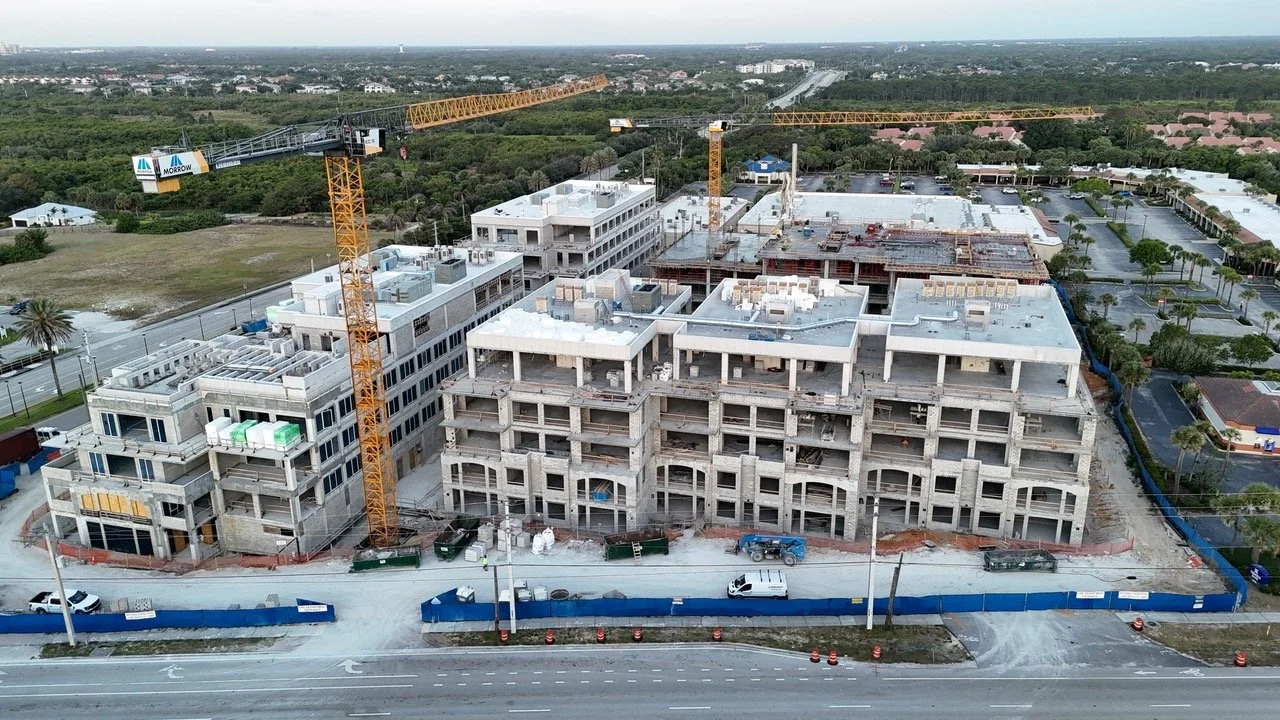 Aerial view of a multi-story building under construction with cranes and construction equipment at a site surrounded by a blue safety fence.