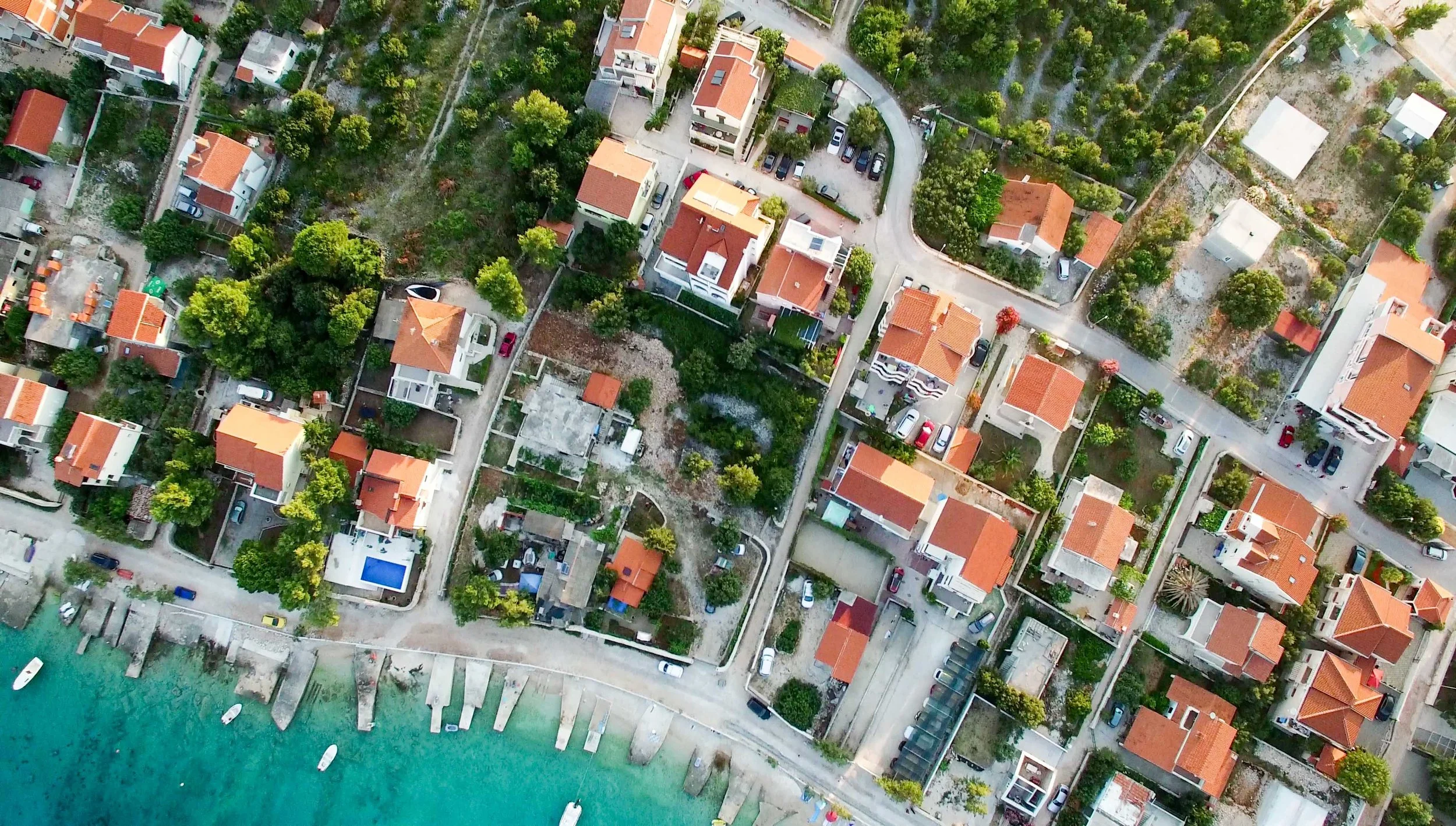 Aerial view of a coastal residential neighborhood with houses, gardens, and trees along the shoreline, with boats docked at the harbor.