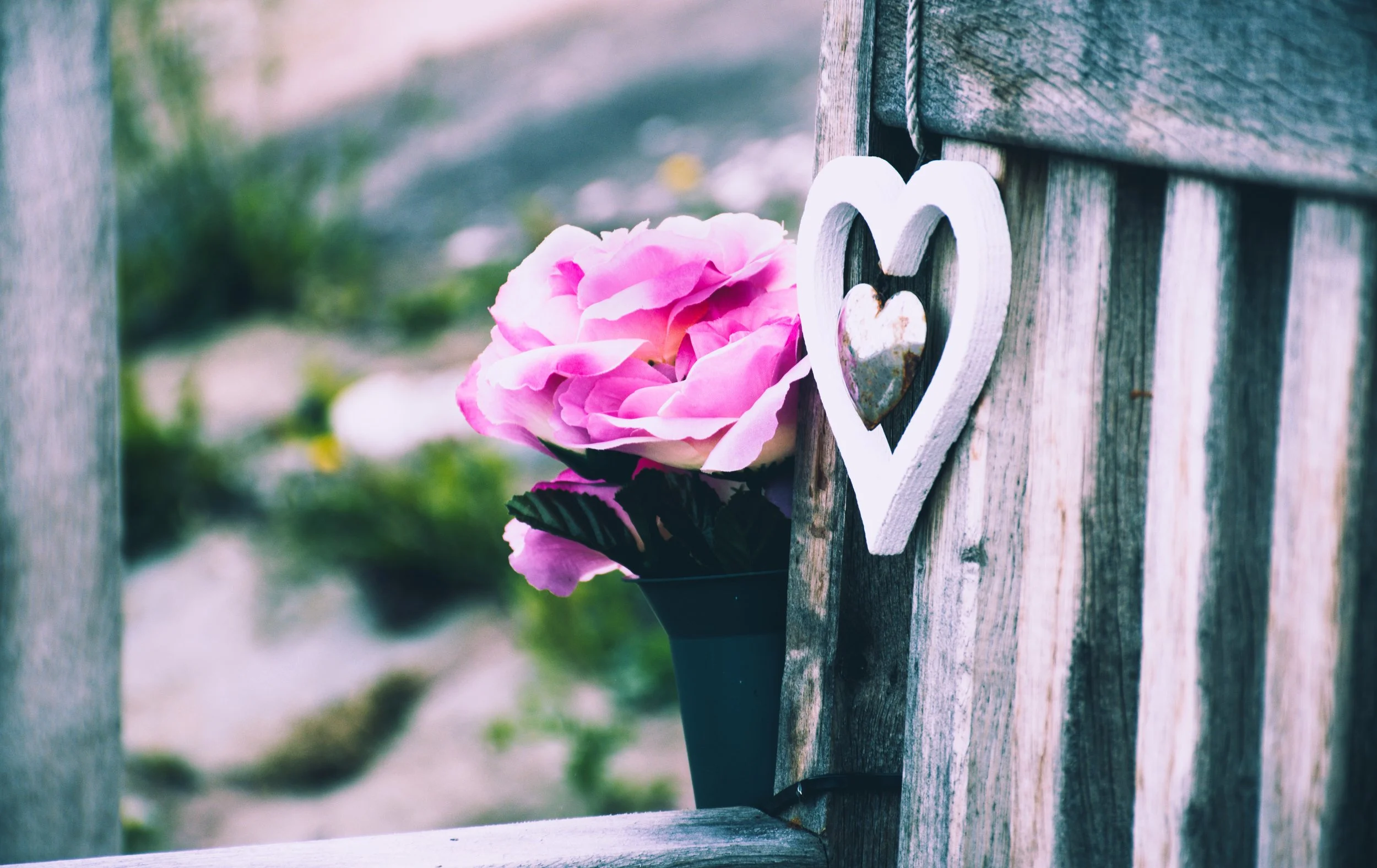 Pink artificial flower in a black vase next to a wooden fence with a white heart decoration.