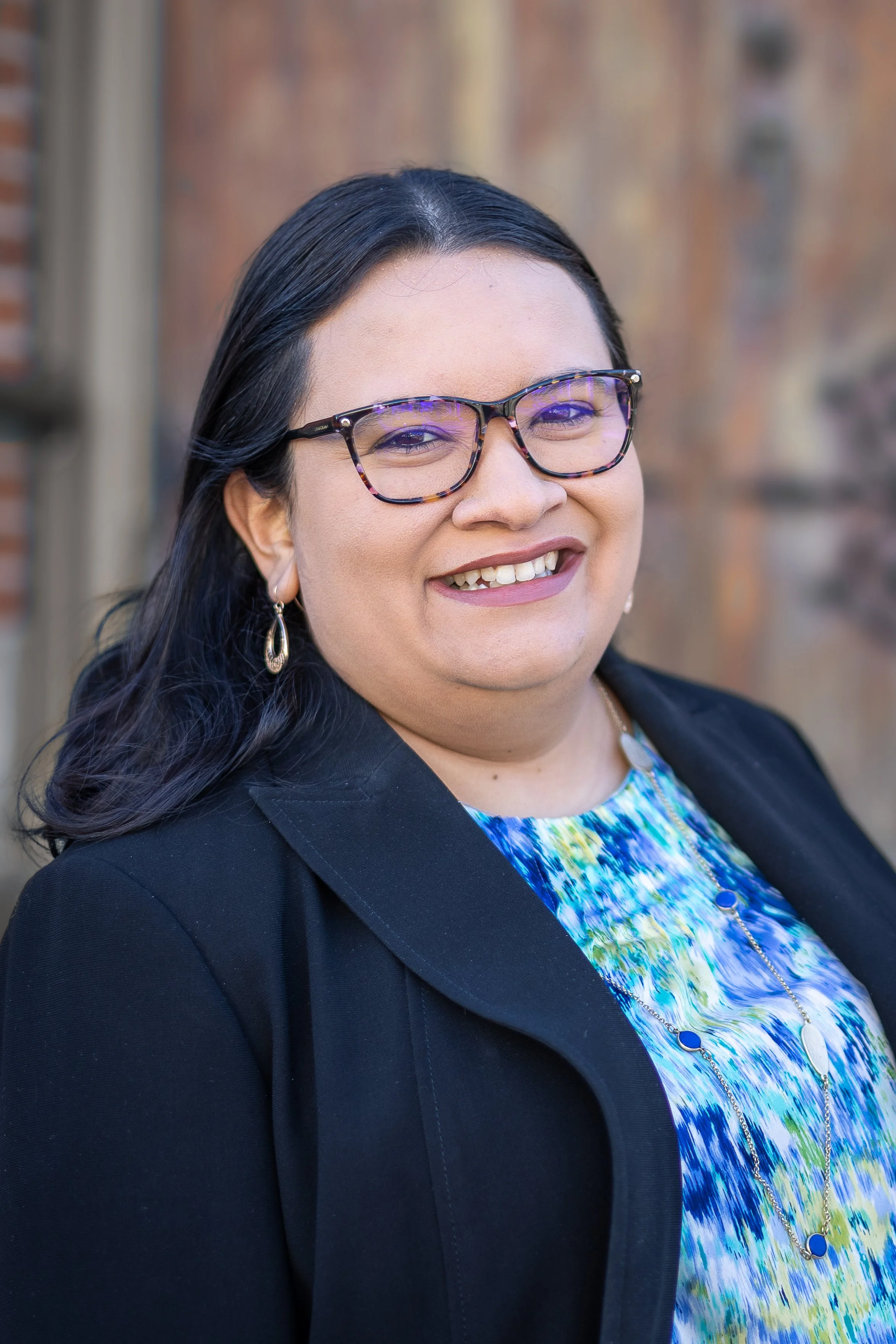 A woman with long dark hair, glasses, and hoop earrings, smiling outdoors against a blurred background of brick and wood.