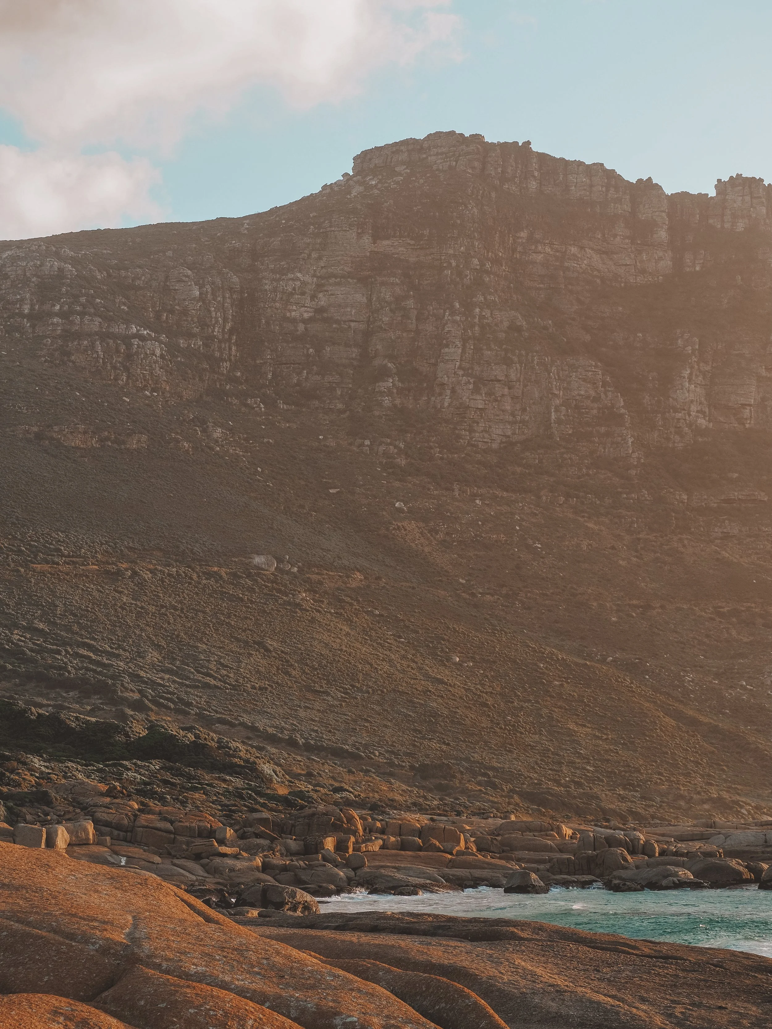 Mountain landscape with rocky shoreline and ocean waves under a partly cloudy sky.