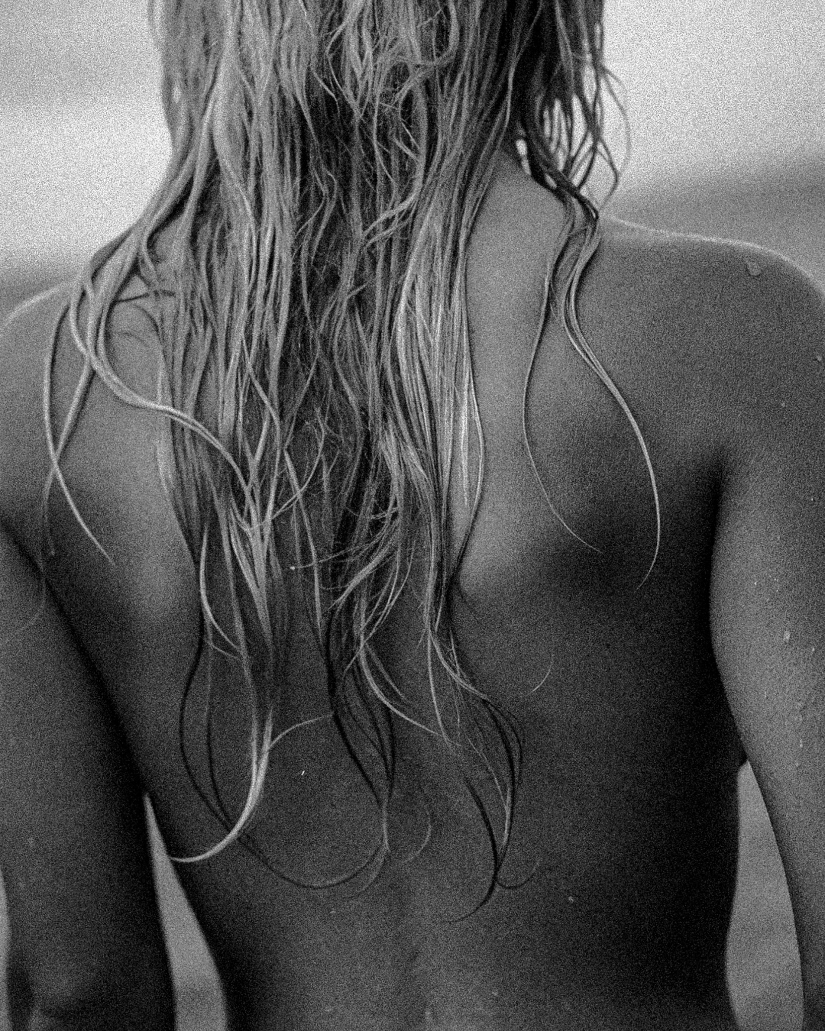 Close-up of a woman's wet, curly hair cascading over her shoulder, with her back and shoulder visible in black and white.