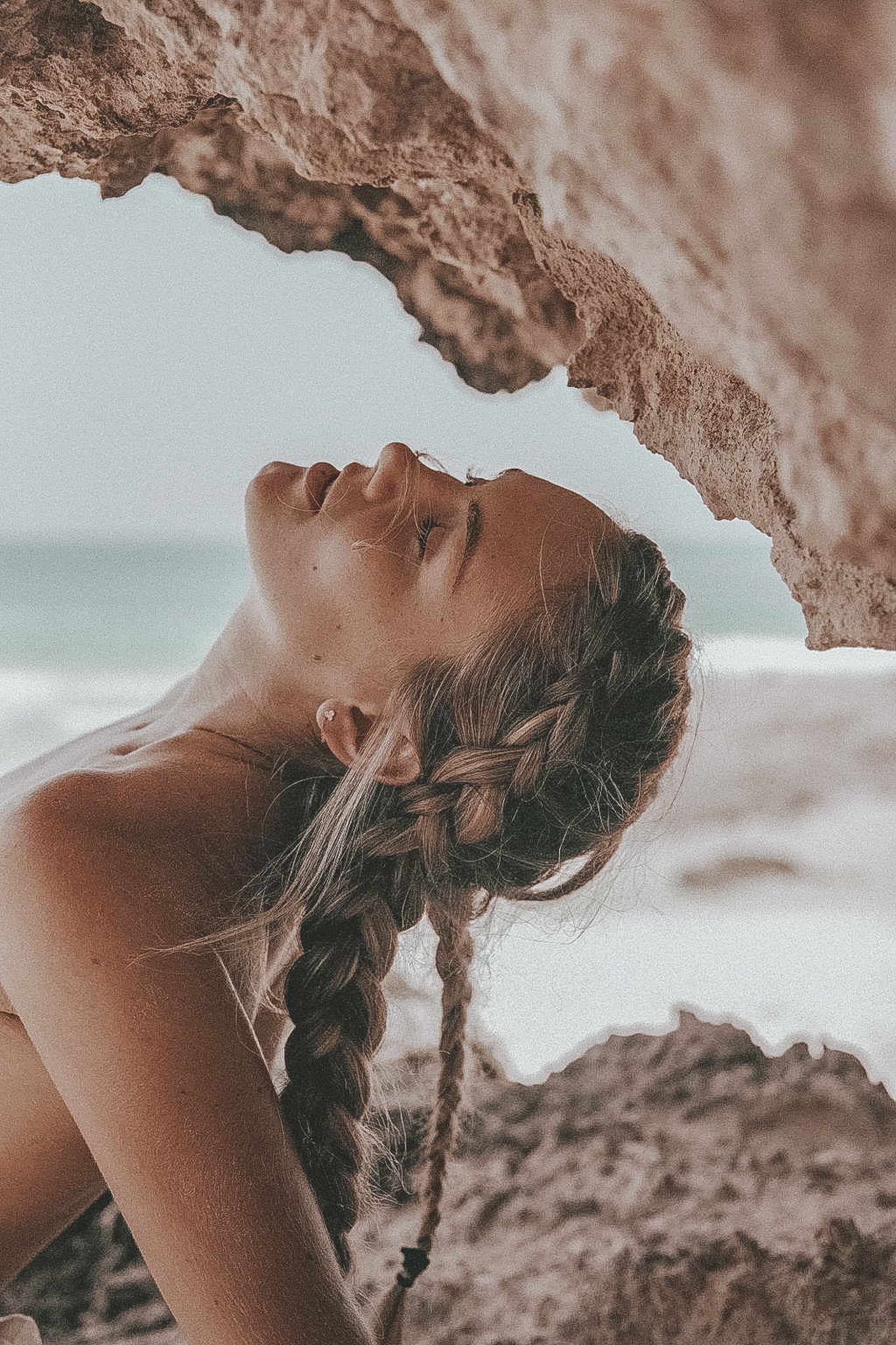 A woman with braids is lying beneath a rocky overhang on the beach, with the ocean visible in the background.