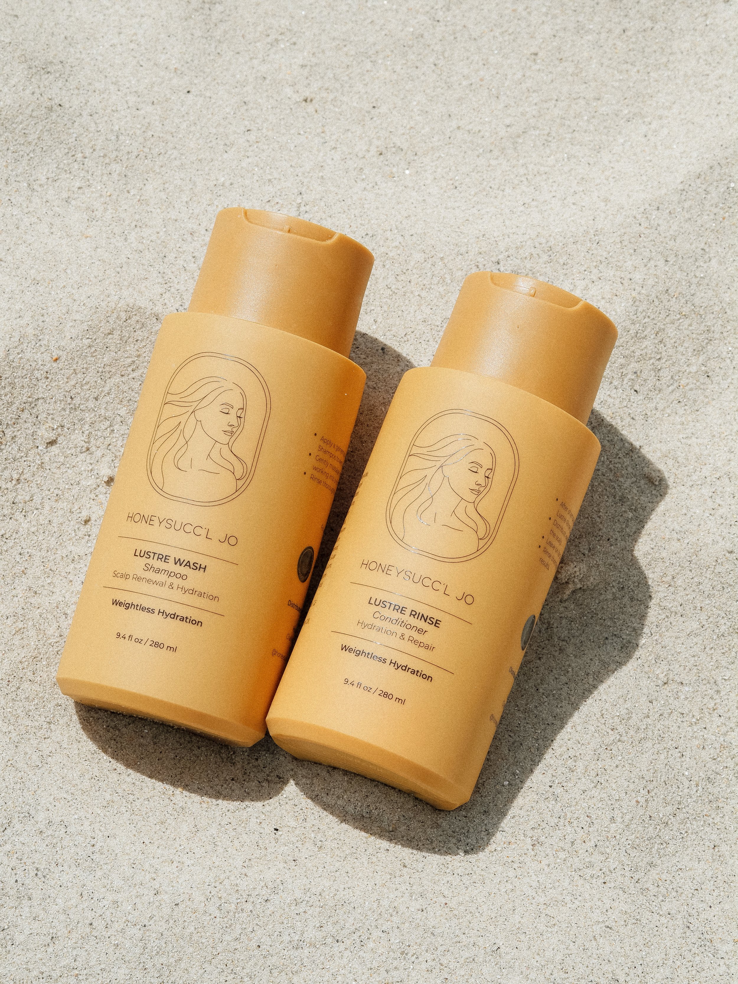 Two bottles of skincare products on sand, one labeled 'Lustre Wash Shampoo' and the other 'Lustre Rinse Conditioner' from Honey Suckle Jo, with a minimalist line drawing of a woman's face on each.