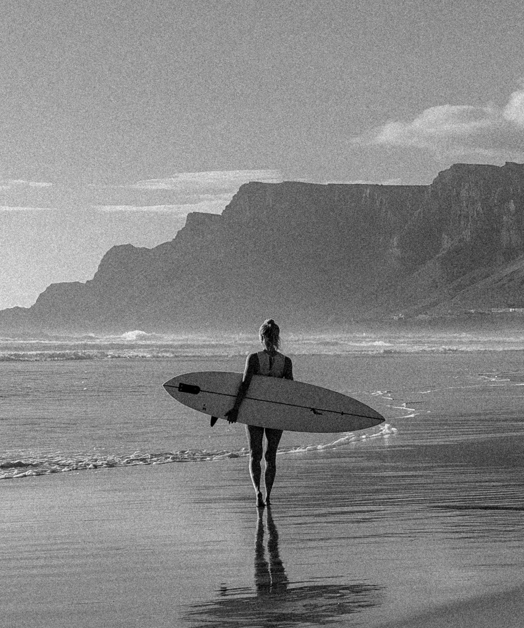 A woman walking on the beach carrying a surfboard, with cliffs in the background and the ocean waves around her, in black and white.