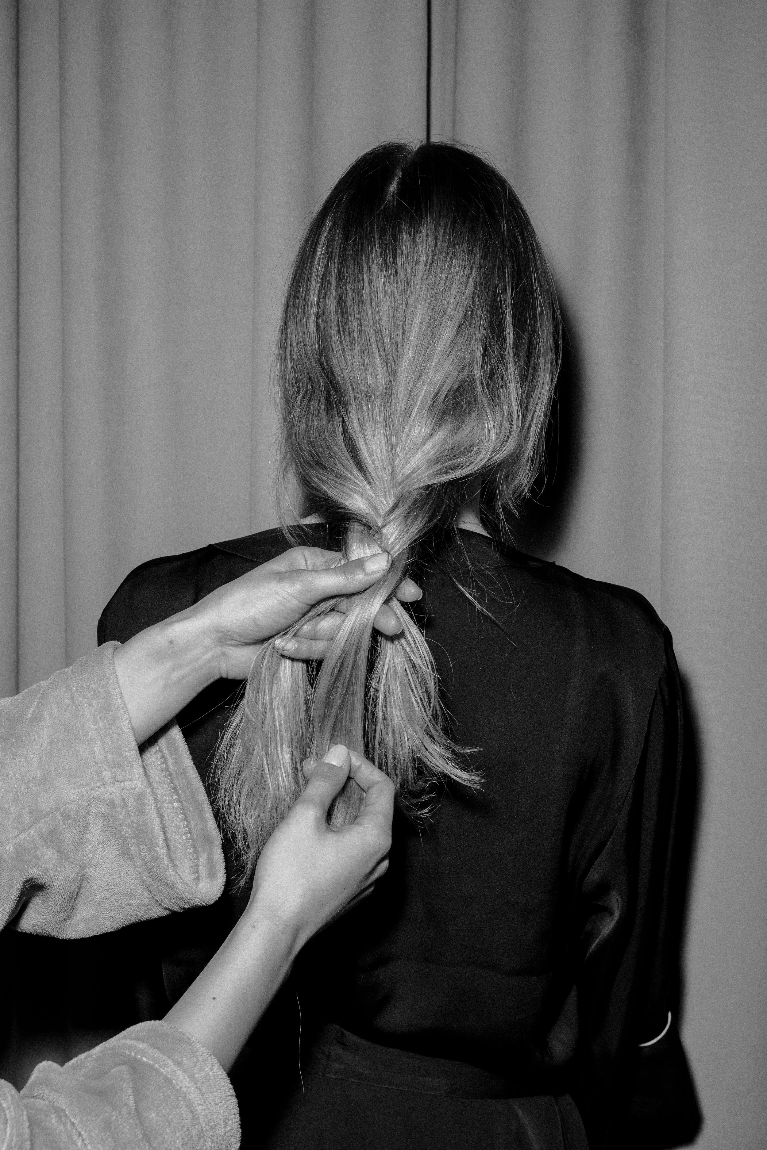 Person with shoulder-length hair having it styled or braided by another person's hands in front of curtain background.