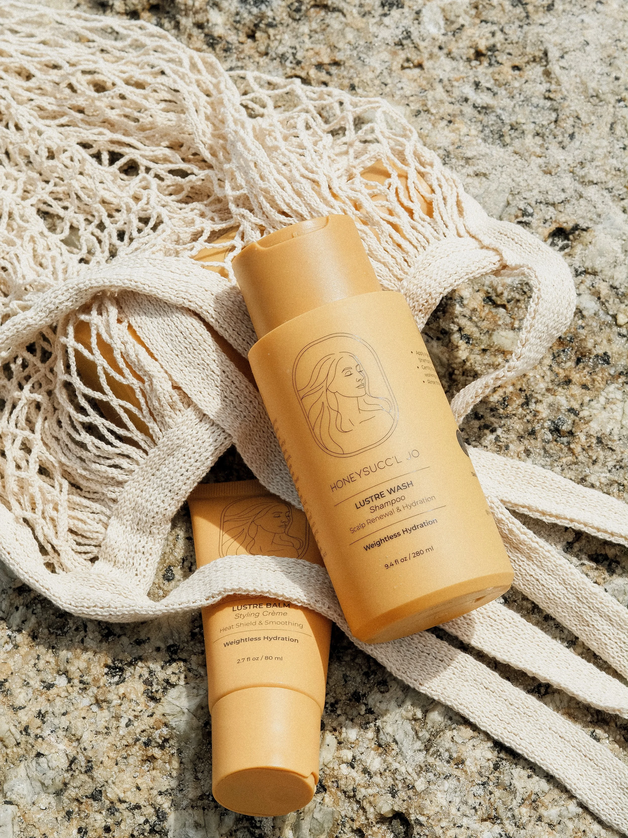 Two bottles of HONEYSUCC'L JO hair products, a shampoo and a styling crème, resting on a beige and brown speckled granite surface, with a white, textured, fringed fabric in the background.