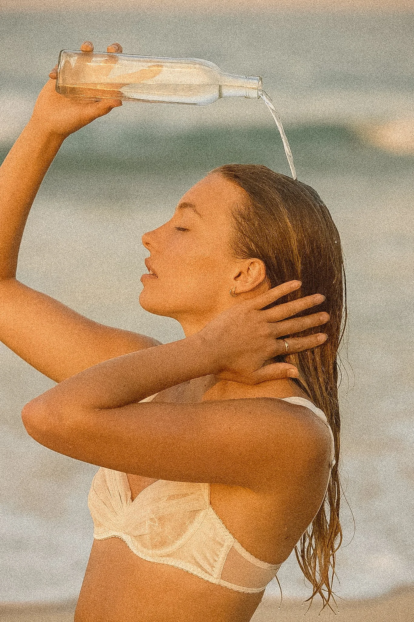 A woman with wet hair is wearing a white top and holding a bottle of water over her head, pouring water onto her hair at the beach.