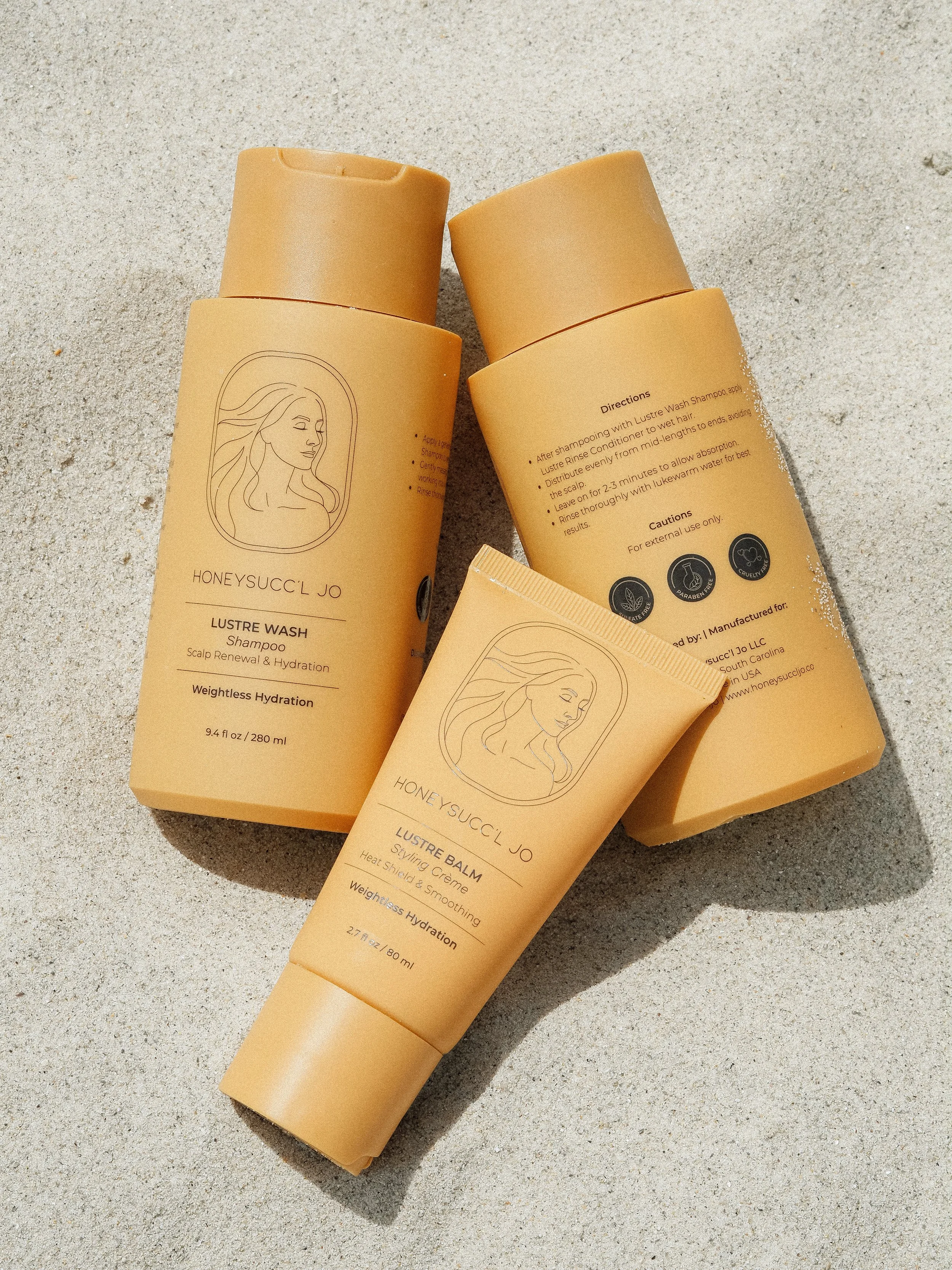 Three skincare product tubes labeled HONEYSUCC'L JO HAIRCARE, positioned on sandy ground, with one tube lying across two upright tubes.
